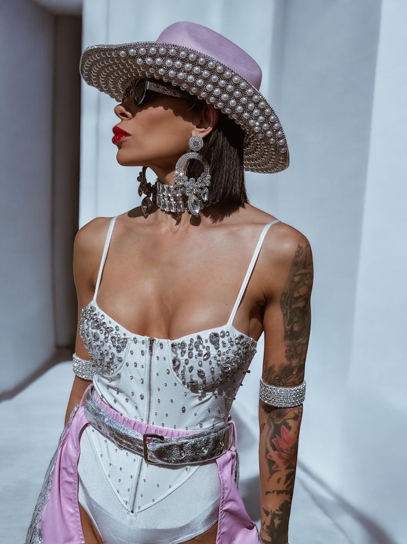 May include: A white bodysuit with a corset style bodice and a silver belt. The bodysuit is covered in rhinestones. The model is wearing a pink cowboy hat with pearl embellishments and large rhinestone earrings.