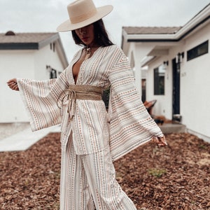 May include: A white and beige patterned kimono with a wide, woven belt. The kimono has long sleeves and a deep V-neck. The model is wearing a wide-brimmed hat and a necklace.