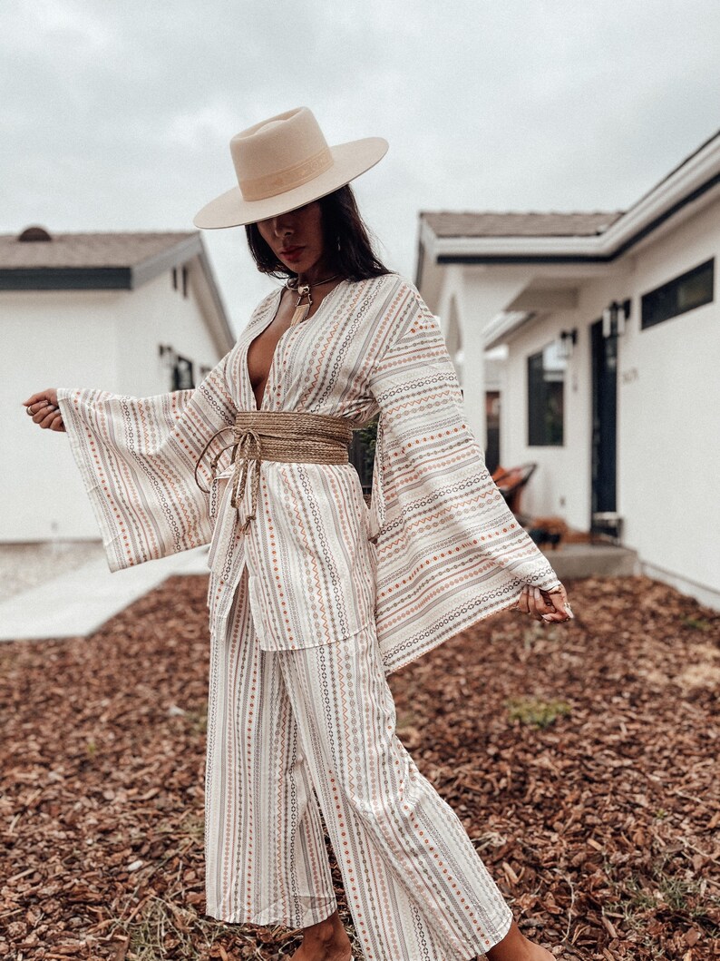 May include: A white and beige patterned kimono with a wide, woven belt. The kimono has long sleeves and a deep V-neck. The model is wearing a wide-brimmed hat and a necklace.