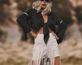 May include: A black cowboy hat with silver embellishments and sunglasses. The outfit includes a black fringed top tied at the waist and off-white fringed shorts. The ensemble evokes a Western theme.
