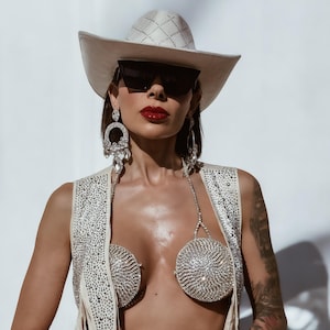 K&ouml;nnte beinhalten: Eine Frau tr&auml;gt einen wei&szlig;en Cowboyhut, eine Sonnenbrille und eine mit Strass besetzte Weste. Sie tr&auml;gt auch einen Strass-BH und Armb&auml;nder.