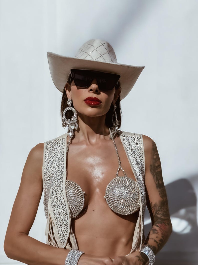 K&ouml;nnte beinhalten: Eine Frau tr&auml;gt einen wei&szlig;en Cowboyhut, eine Sonnenbrille und eine mit Strass besetzte Weste. Sie tr&auml;gt auch einen Strass-BH und Armb&auml;nder.