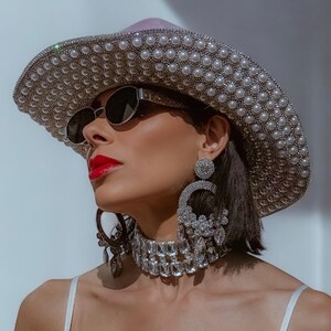 May include: A woman wearing a large, silver and pearl embellished cowboy hat, oversized sunglasses, and a crystal choker necklace.