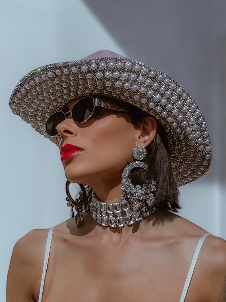 May include: A woman wearing a large, silver and pearl embellished cowboy hat, oversized sunglasses, and a crystal choker necklace.