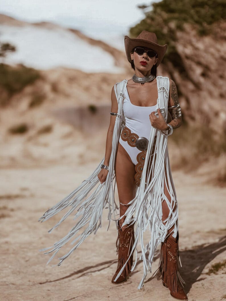 May include: A white swimsuit and fringed vest, complemented by a brown cowboy hat and boots. The outfit is accessorized with silver jewellery. The boots have a brown leather finish. The overall look is a blend of bohemian and Western styles.