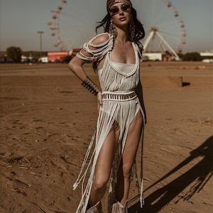 Puede incluir: Una mujer con un atuendo crema con flecos, pa&ntilde;uelo a juego y gafas de sol. El atuendo incluye un top, una falda y botas con flecos. El fondo muestra una noria y un paisaje des&eacute;rtico.