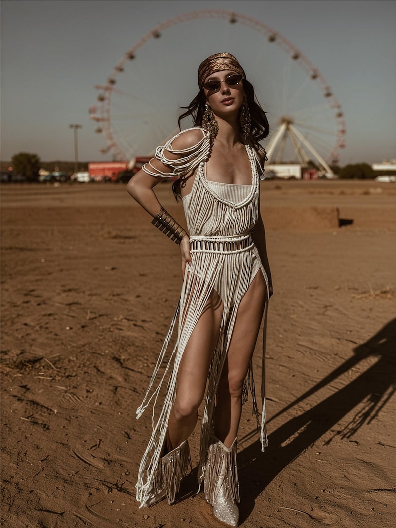 Puede incluir: Una mujer con un atuendo crema con flecos, pa&ntilde;uelo a juego y gafas de sol. El atuendo incluye un top, una falda y botas con flecos. El fondo muestra una noria y un paisaje des&eacute;rtico.
