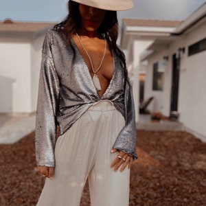 May include: A woman wearing a silver sequined top with a deep V-neckline and long sleeves. She is also wearing a white hat and a white necklace with a tassel pendant. The woman is wearing white pants.
