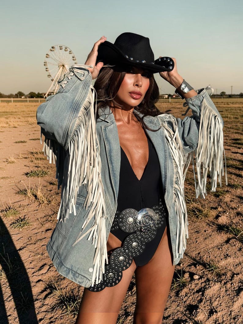 May include: A black cowboy hat with silver studs, a light blue denim jacket featuring long white fringe on the sleeves and shoulders, a black bodysuit, and a wide black leather belt adorned with large circular silver concho-style metal plates.
