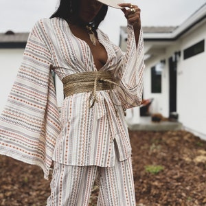 May include: A white and brown striped pantsuit with a wide, woven belt. The pantsuit has a deep V-neckline and long, wide sleeves. The person is wearing a wide-brimmed hat.