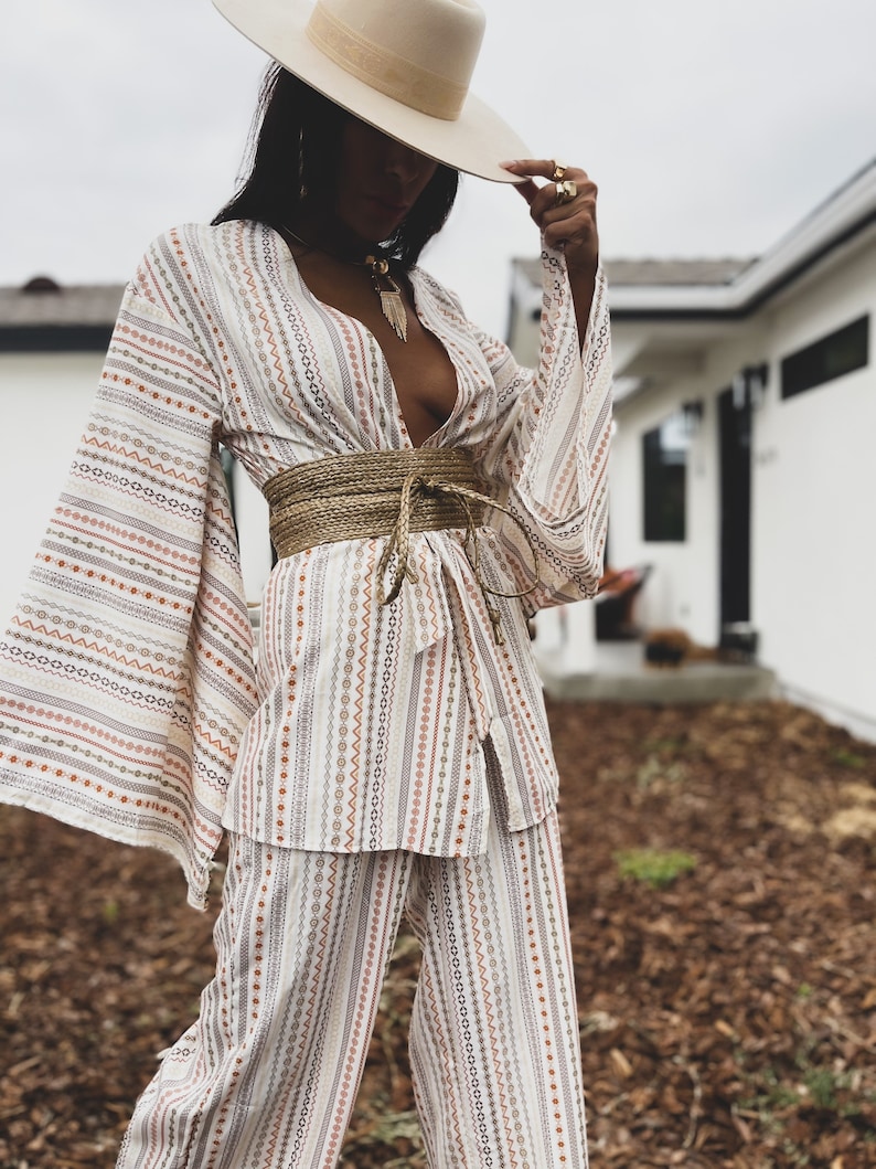 May include: A white and brown striped pantsuit with a wide, woven belt. The pantsuit has a deep V-neckline and long, wide sleeves. The person is wearing a wide-brimmed hat.