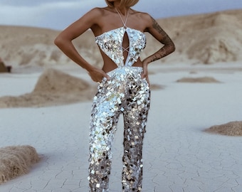 Peut inclure: Combinaison argentée à paillettes avec un col halter et des découpes latérales. La combinaison présente une coupe droite et est associée à des talons argentés à bout pointu. La tenue est réfléchissante et accrocheuse, parfaite pour un événement spécial.