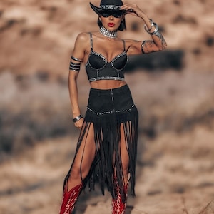 May include: A black cowboy hat with silver embellishments and sunglasses. The outfit includes a black bra top with silver studs, a black skirt with fringe, and red cowboy boots with white stitching. The background is a desert landscape.
