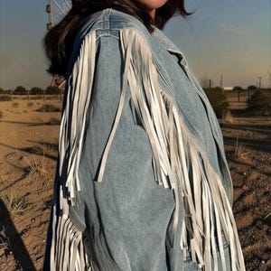 May include: A light blue denim jacket featuring long white fringe detailing along the shoulders and sleeves. The jacket has a classic collar and a button-down front, with the fringe extending from the shoulder seam down the length of the sleeve.