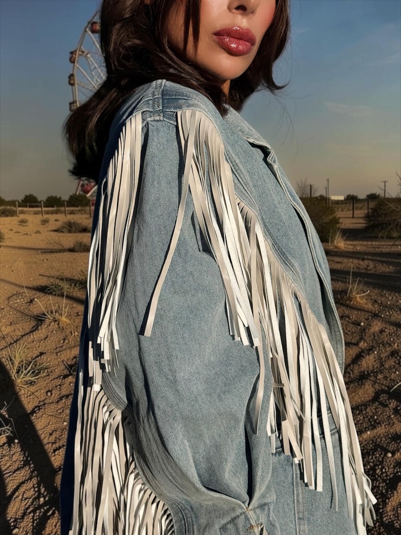 May include: A light blue denim jacket featuring long white fringe detailing along the shoulders and sleeves. The jacket has a classic collar and a button-down front, with the fringe extending from the shoulder seam down the length of the sleeve.