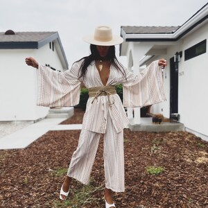 May include: A white and pink striped wide-leg pantsuit with a wide, flowing kimono-style top. The outfit is cinched at the waist with a brown rope belt. The model is wearing a white hat and white heels.