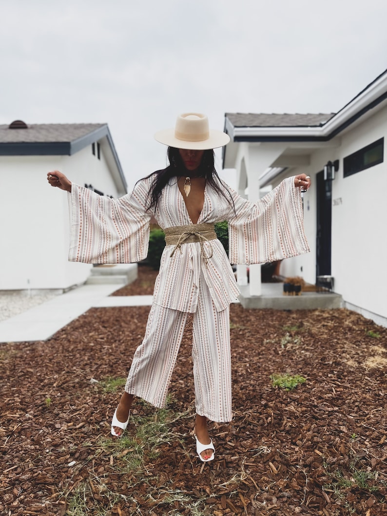 May include: A white and pink striped wide-leg pantsuit with a wide, flowing kimono-style top. The outfit is cinched at the waist with a brown rope belt. The model is wearing a white hat and white heels.