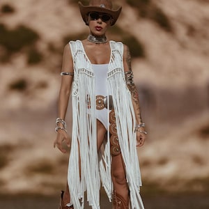 May include: A woman in a brown cowboy hat and sunglasses, wearing a white fringed vest and bodysuit. She is also wearing a silver belt and brown cowboy boots. The background is a blurred desert landscape.