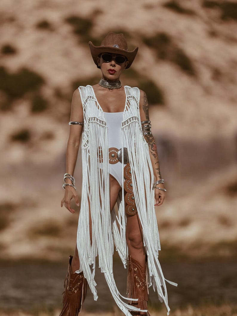 May include: A woman in a brown cowboy hat and sunglasses, wearing a white fringed vest and bodysuit. She is also wearing a silver belt and brown cowboy boots. The background is a blurred desert landscape.
