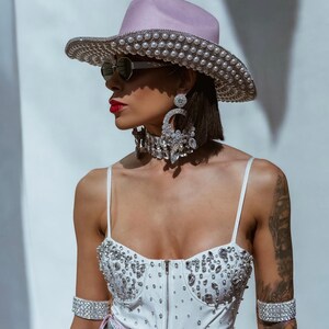 May include: A woman wearing a white bodysuit with rhinestones, a pink cowboy hat with pearl embellishments, and silver jewelry. She is also wearing sunglasses and has a tattoo on her arm.