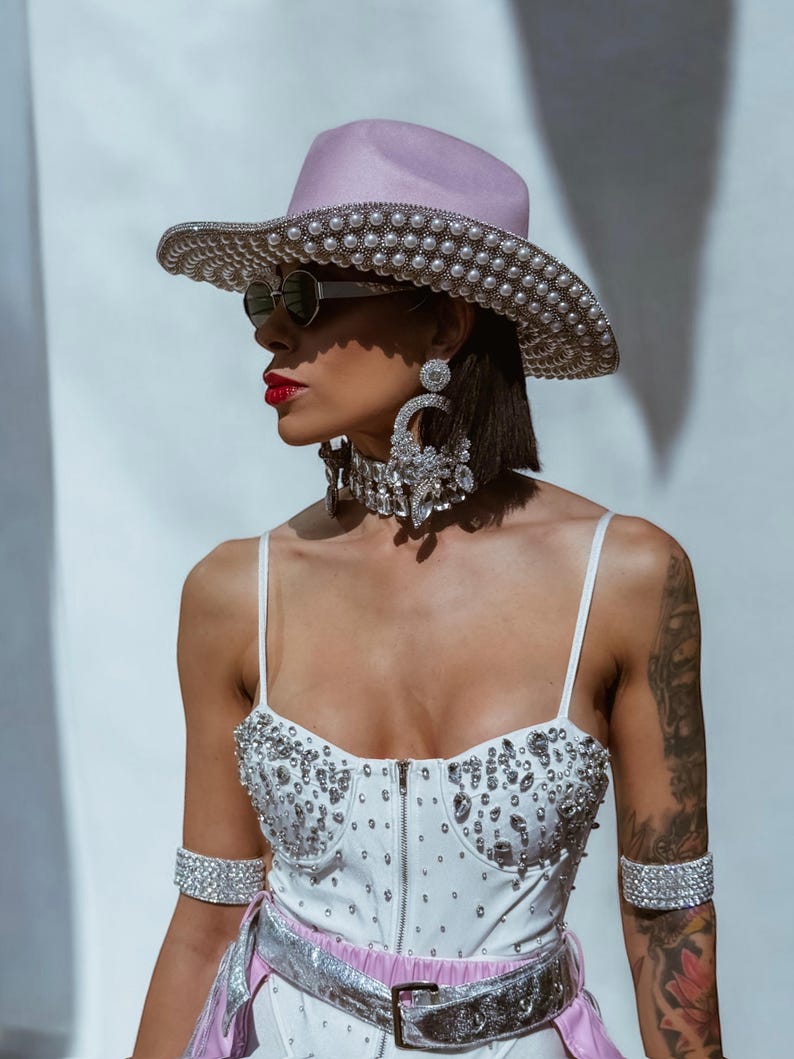 May include: A woman wearing a white bodysuit with rhinestones, a pink cowboy hat with pearl embellishments, and silver jewelry. She is also wearing sunglasses and has a tattoo on her arm.