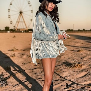 May include: A light blue denim jacket featuring long white fringe detailing along the sleeves and back, paired with a black cowboy hat and black boots. The jacket includes lace-up side accents with crisscross stitching on the sleeves.