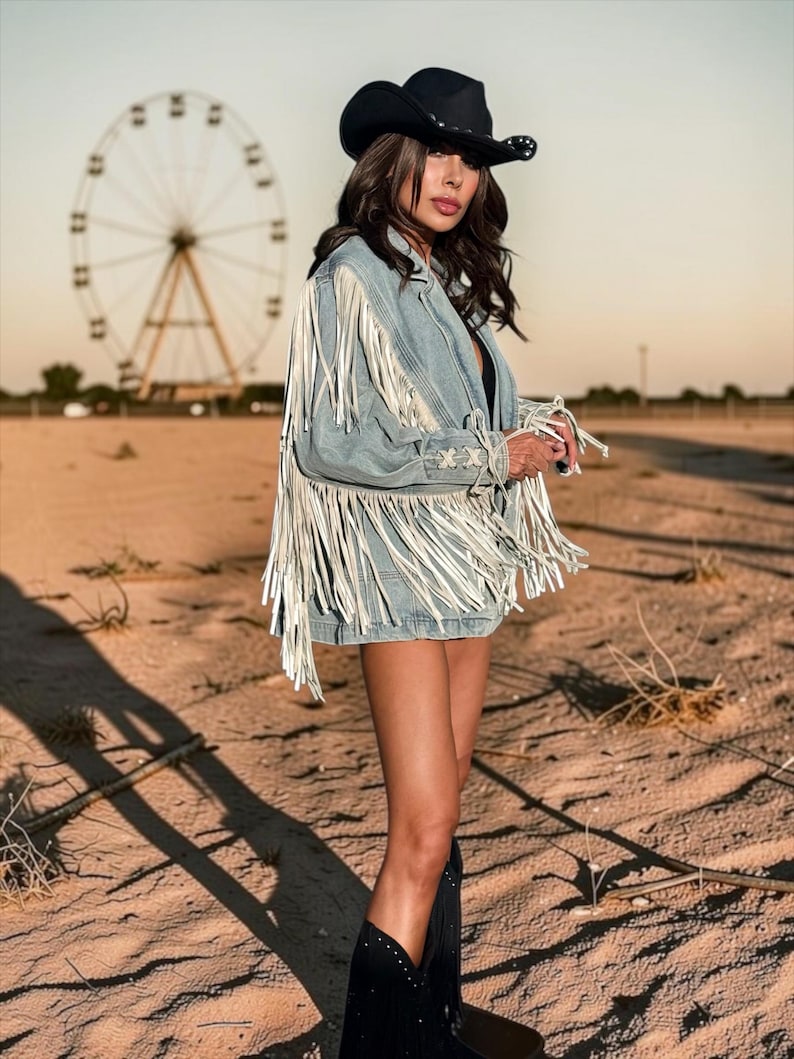 May include: A light blue denim jacket featuring long white fringe detailing along the sleeves and back, paired with a black cowboy hat and black boots. The jacket includes lace-up side accents with crisscross stitching on the sleeves.