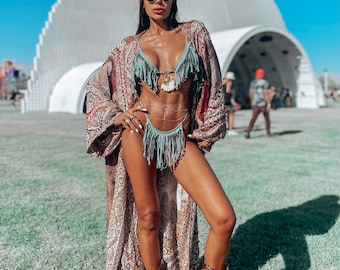 May include: A woman wearing a green fringe bikini top and bottom, a brown paisley kimono, a red bandana, and brown cowboy boots. She is standing in a grassy field in front of a large white structure.