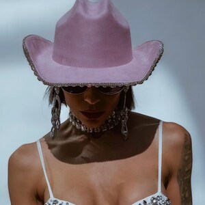 May include: A pink cowboy hat with a rhinestone trim. The hat is worn by a person with a white top with rhinestones and silver jewelry.