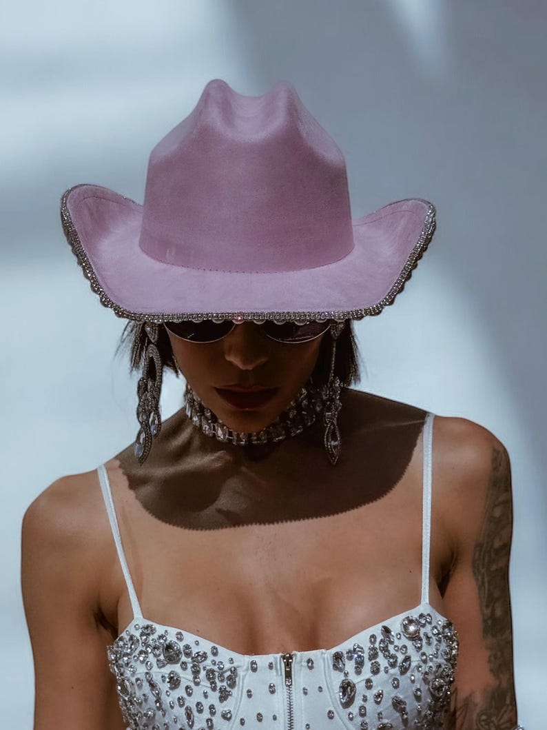 May include: A pink cowboy hat with a rhinestone trim. The hat is worn by a person with a white top with rhinestones and silver jewelry.