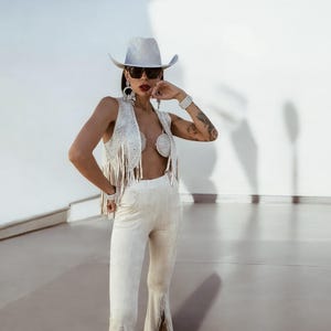K&ouml;nnte beinhalten: Ein wei&szlig;er Cowboyhut mit einem hellblauen Band, wei&szlig;e Sonnenbrille, eine wei&szlig;e Fransenweste mit Perlenmuster und eine wei&szlig;e Hose mit Fransen.