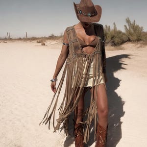 May include: A brown suede cowboy hat with turquoise detailing and a long fringe vest. The vest is worn with a matching skirt and brown cowboy boots with fringe. The outfit is set in a desert landscape.
