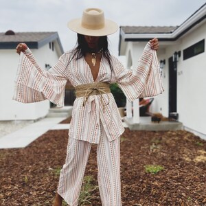 May include: A woman wearing a white and brown striped pantsuit with a wide, brown woven belt. She is wearing a wide-brimmed, cream-colored hat and white shoes.