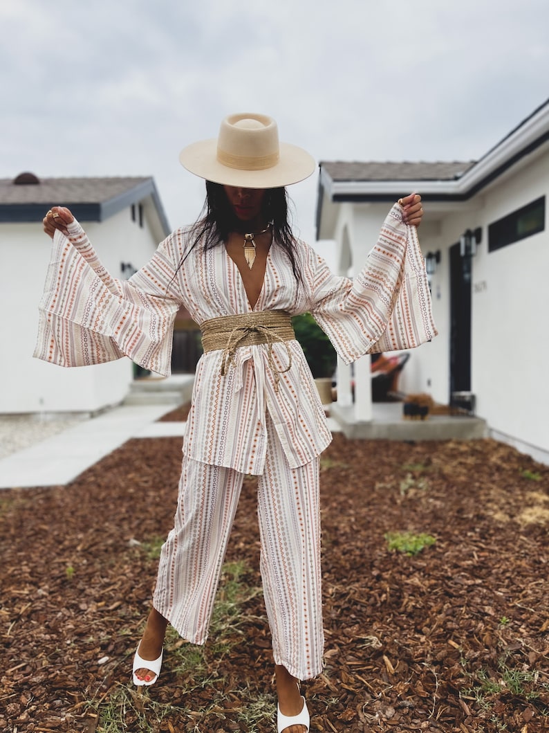May include: A woman wearing a white and brown striped pantsuit with a wide, brown woven belt. She is wearing a wide-brimmed, cream-colored hat and white shoes.