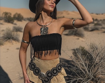 May include: A woman wearing a black cowboy hat, black fringed tube top, and a tan skirt with a decorative black belt. She is also wearing a silver necklace and arm cuffs. The background is a desert landscape with sand and sparse vegetation.
