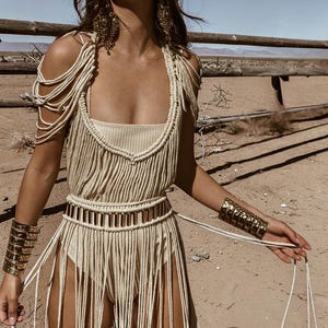 Puede incluir: Vestido de macram&eacute; beige con flecos, escote cuadrado y cintur&oacute;n a juego. El vestido presenta tirantes y se complementa con brazaletes dorados. El fondo es un paisaje des&eacute;rtico.
