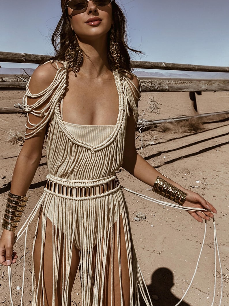 Puede incluir: Vestido de macram&eacute; beige con flecos, escote cuadrado y cintur&oacute;n a juego. El vestido presenta tirantes y se complementa con brazaletes dorados. El fondo es un paisaje des&eacute;rtico.