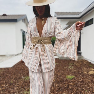 May include: A white and brown striped pantsuit with a wide, brown woven belt. The pantsuit has a deep V-neckline and long, wide sleeves. The person is wearing a wide-brimmed, cream-colored hat.