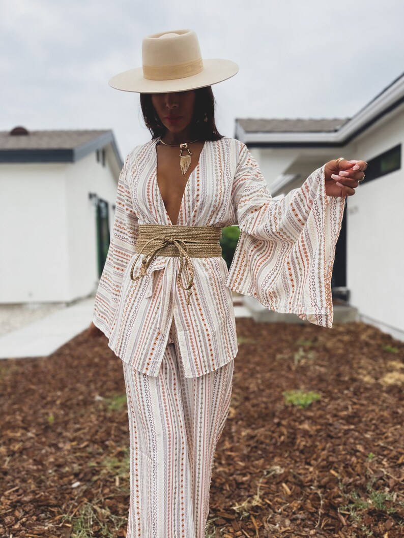May include: A white and brown striped pantsuit with a wide, brown woven belt. The pantsuit has a deep V-neckline and long, wide sleeves. The person is wearing a wide-brimmed, cream-colored hat.