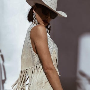 K&ouml;nnte beinhalten: Eine Frau tr&auml;gt einen wei&szlig;en Cowboyhut, eine Sonnenbrille und eine beige Wildlederweste mit Fransen und Strasssteinen. Die Weste ist &auml;rmellos und hat eine figurbetonte Silhouette.