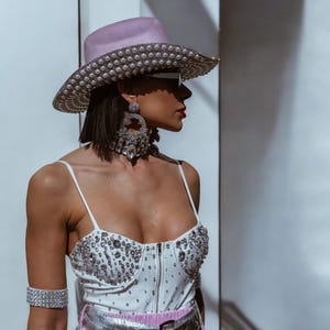 May include: A woman wearing a white bodysuit with rhinestones, a pink cowboy hat with pearl embellishments, a silver belt, and large crystal earrings.