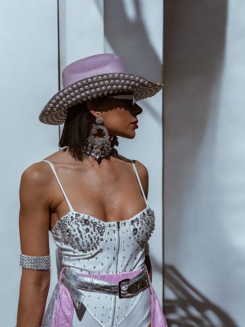 May include: A woman wearing a white bodysuit with rhinestones, a pink cowboy hat with pearl embellishments, a silver belt, and large crystal earrings.