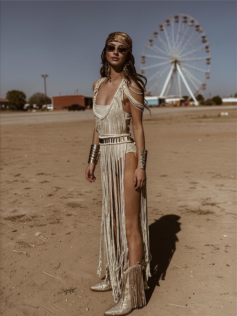 Puede incluir: Una mujer con un vestido crema con flecos, brazaletes dorados y botas con flecos frente a una noria. Tambi&eacute;n lleva un pa&ntilde;uelo dorado en la cabeza y gafas de sol. El atuendo es de estilo bohemio.