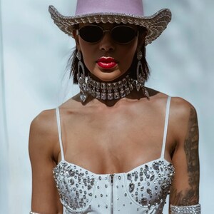 May include: A woman wearing a pink cowboy hat with rhinestones, a white rhinestone-studded top, and large rhinestone earrings. She is also wearing a rhinestone choker and bracelets.