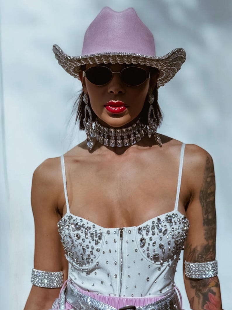 May include: A woman wearing a pink cowboy hat with rhinestones, a white rhinestone-studded top, and large rhinestone earrings. She is also wearing a rhinestone choker and bracelets.