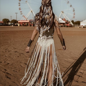 Puede incluir: Una persona con un atuendo bohemio se aleja de la c&aacute;mara. Lleva una falda con flecos color crema, un top a juego y un pa&ntilde;uelo en la cabeza estampado adornado con plumas. Brazaletes dorados y una noria en el fondo completan el look festivalero.