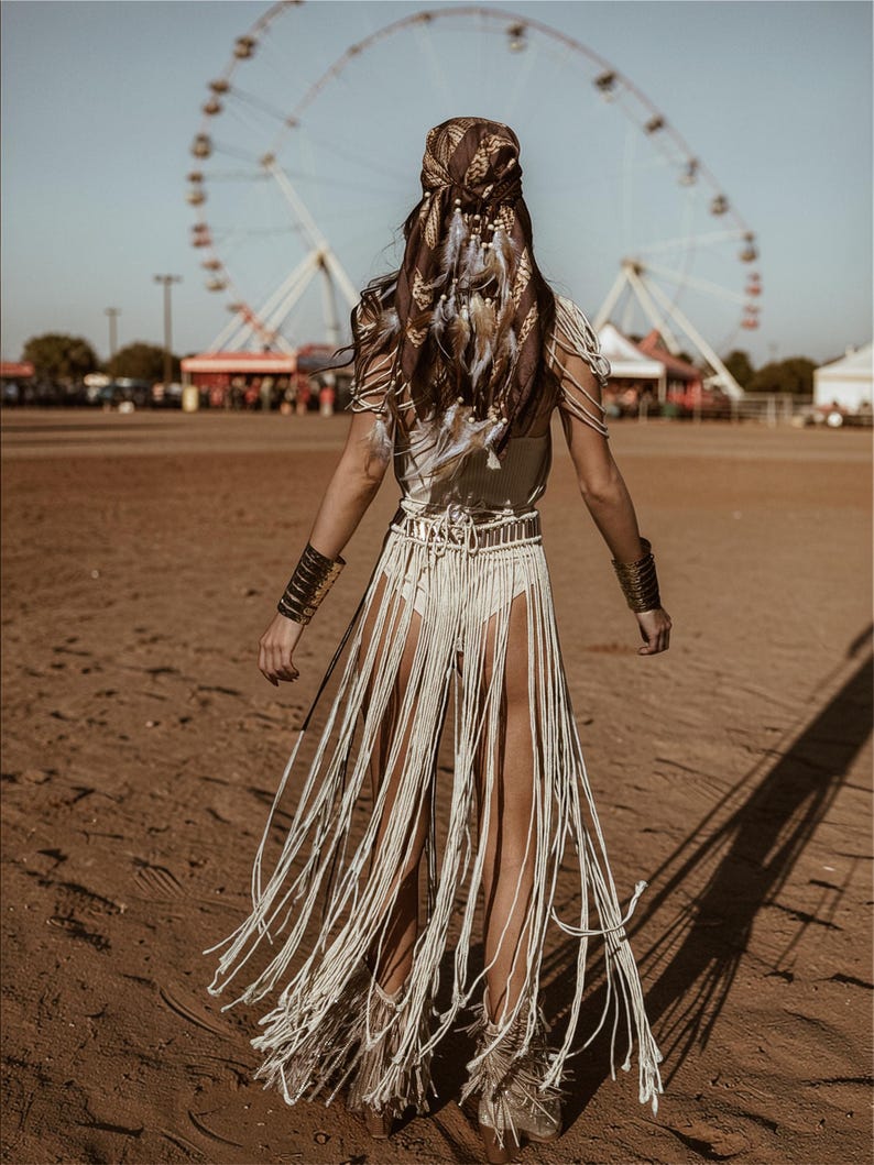 Puede incluir: Una persona con un atuendo bohemio se aleja de la c&aacute;mara. Lleva una falda con flecos color crema, un top a juego y un pa&ntilde;uelo en la cabeza estampado adornado con plumas. Brazaletes dorados y una noria en el fondo completan el look festivalero.