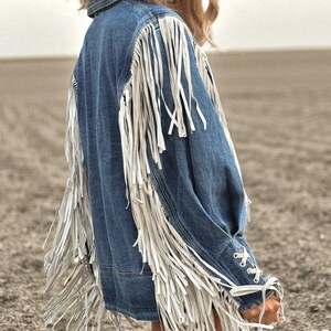 May include: A denim jacket with long white fringe detailing on the sleeves and back. The jacket has a classic collar and button closure. The sleeves are laced at the wrist. The jacket is a medium wash blue.