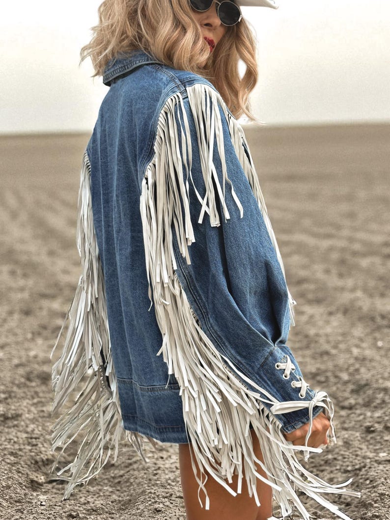 May include: A denim jacket with long white fringe detailing on the sleeves and back. The jacket has a classic collar and button closure. The sleeves are laced at the wrist. The jacket is a medium wash blue.