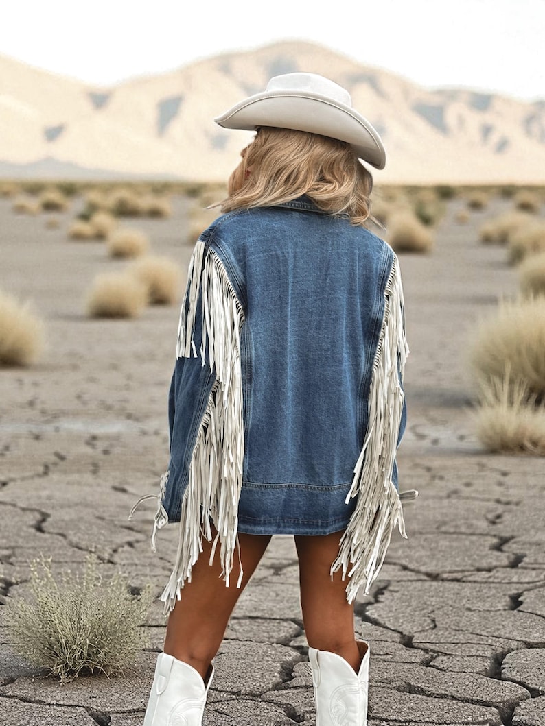 May include: A denim jacket with white fringe detailing and a white cowboy hat. The jacket is a medium wash blue and the fringe hangs from the sleeves and back. The person is wearing white cowboy boots.